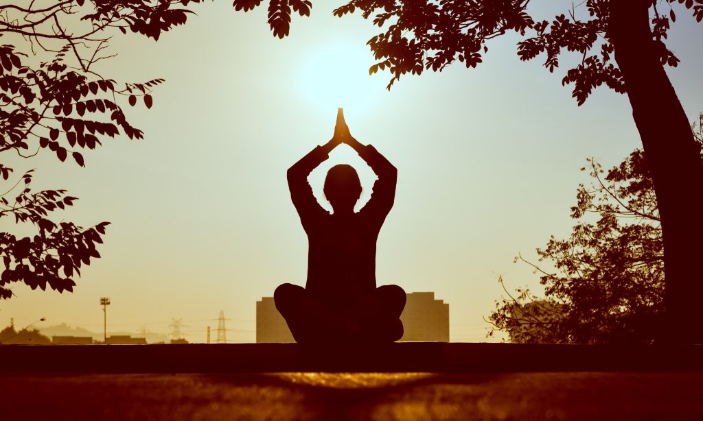 Shadow figure of a girl sitting under a tree facing the sun and meditating with her hands in prayer. There are many benefits of meditation for physical and mental well-being.