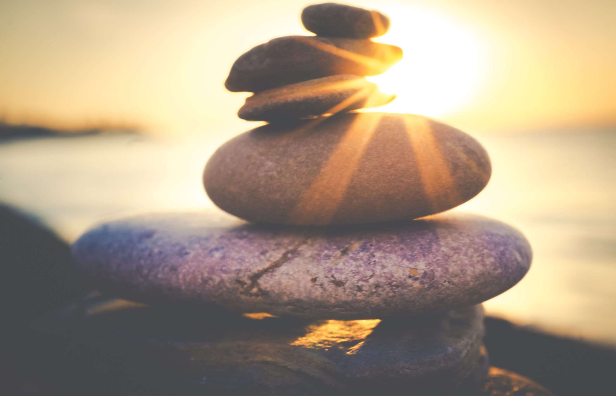 Image showing a pile of rocks by the ocean with the sun in the background to demonstrate serenity. Meditation has so many great benefits, such as improved emotional well-being and boosted immune function.