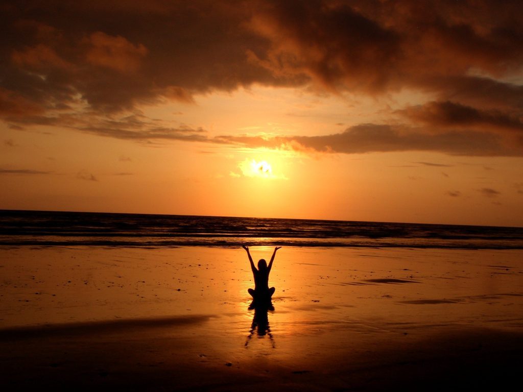 Sunset on a beach with a girl sitting on the beach cross-legged and reaching her arms up to the sky, in e meditative kind of way.