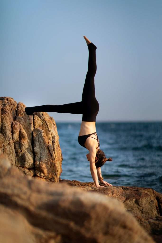 Woman doing a supported handstand by a rock at the sea. One leg is resting on the rock, the other lifting up. Yoga inversions like handstand are a great way to energise the body.